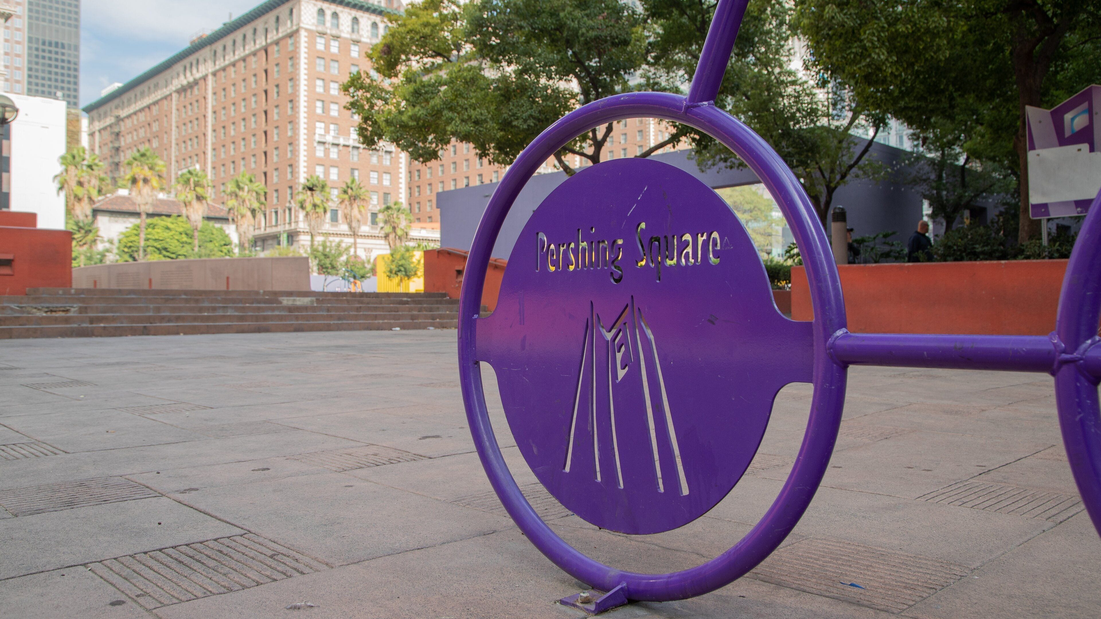 Un letrero púrpura con la inscripción 'Pershing Square', frente a un paisaje urbano con edificios y árboles.