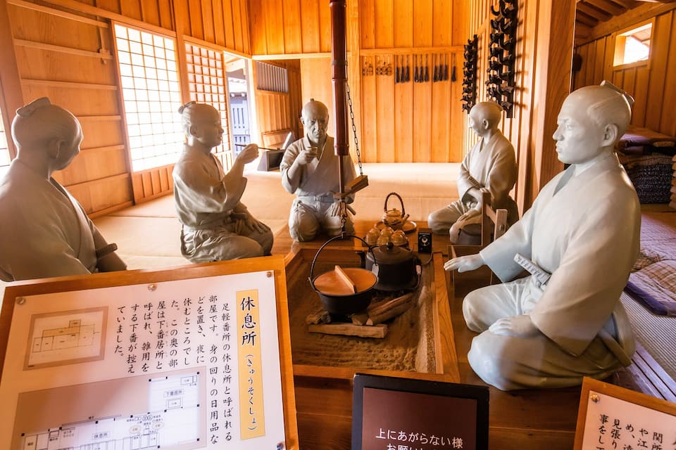A traditional Japanese room with tatami flooring, wooden walls, and a tatami mat bed. There are several statues of historical figures and a tea set on a low table.