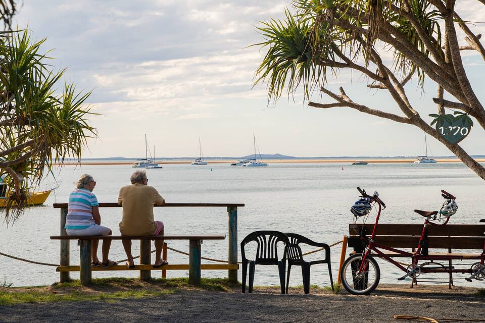 Two people sitting on a bench overlooking a calm body of water with boats and a beach in the distance.