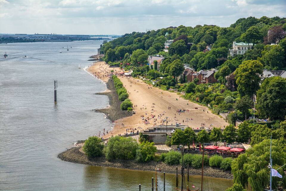 A riverside beach with people, a bridge in the distance, and houses on a hillside.