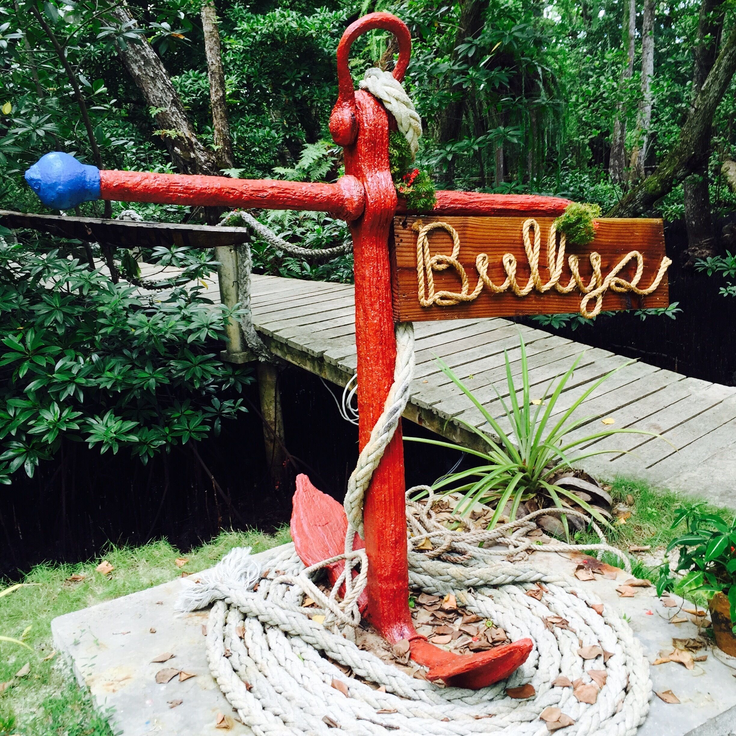 A red hand pump with a rope wrapped around it, a wooden sign with the word "Bullya," and a wooden bridge in a lush green environment.