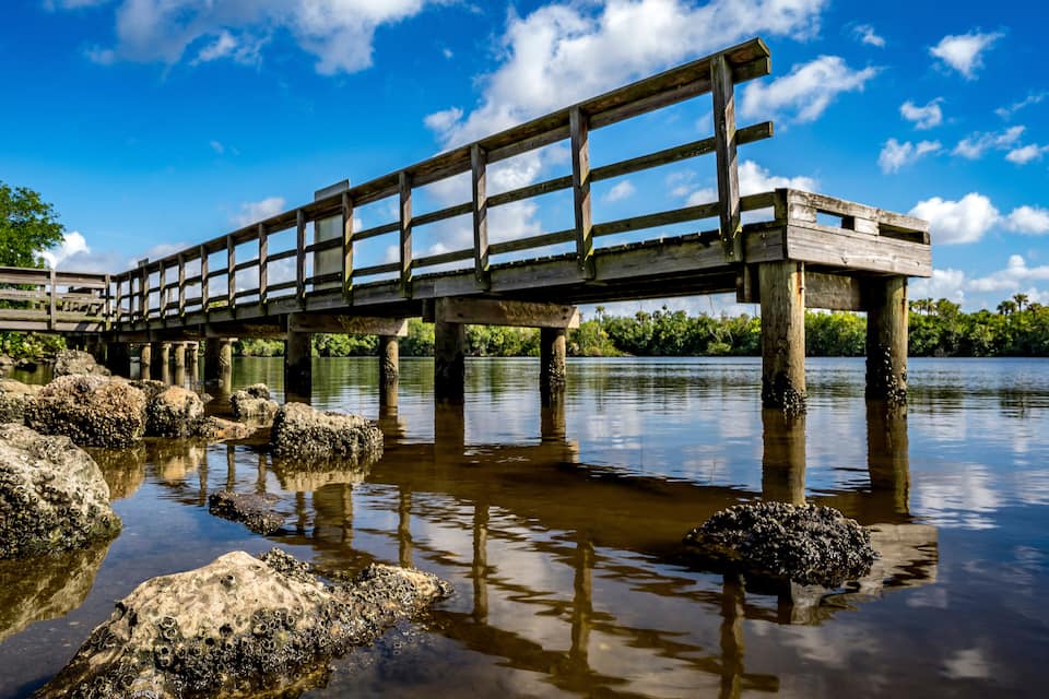 A wooden bridge over a calm body of water with rocks in the foreground and trees in the background.