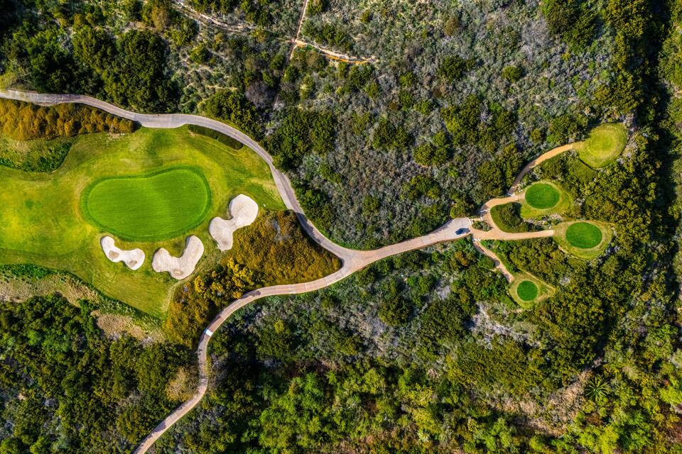 An aerial view of a golf course with a winding road, sand bunkers, and lush greenery.