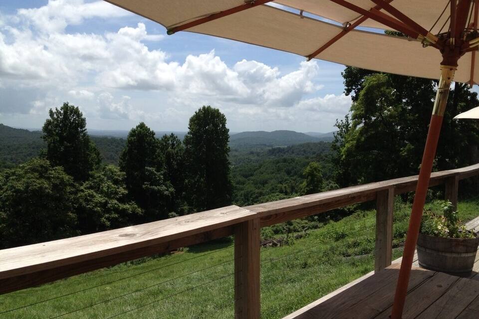 Terraza de madera con un paraguas color beige que da a un paisaje verde exuberante con árboles y montañas a lo lejos.