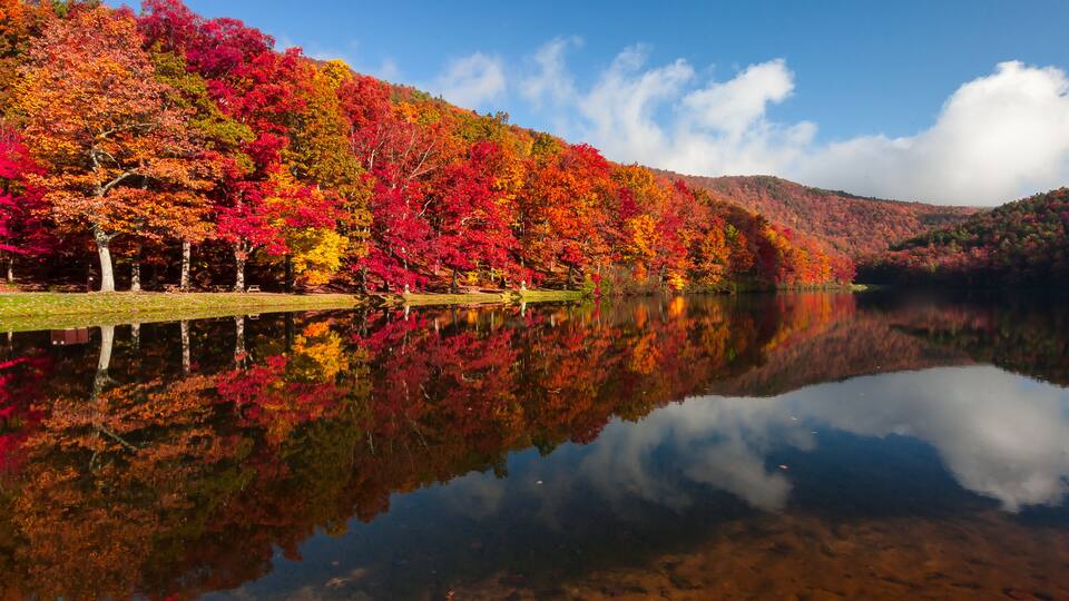 A lake reflecting colorful autumn trees, a clear sky, and a hilly landscape.