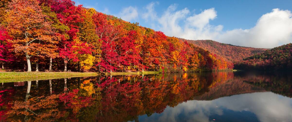 A lake reflecting colorful autumn trees, a clear sky, and a hilly landscape.