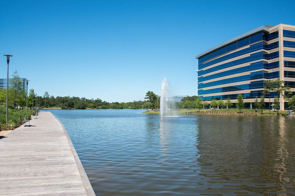 A calm lake with a fountain, a wooden walkway, and modern buildings in the background.