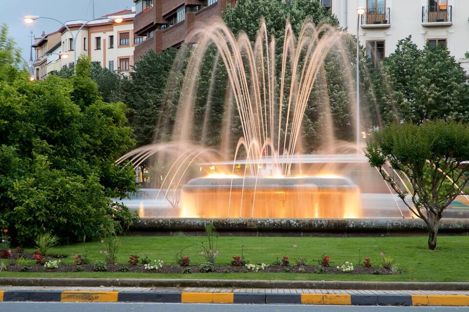 Een fontein met waterstralen in een park, met bomen en gebouwen op de achtergrond.
