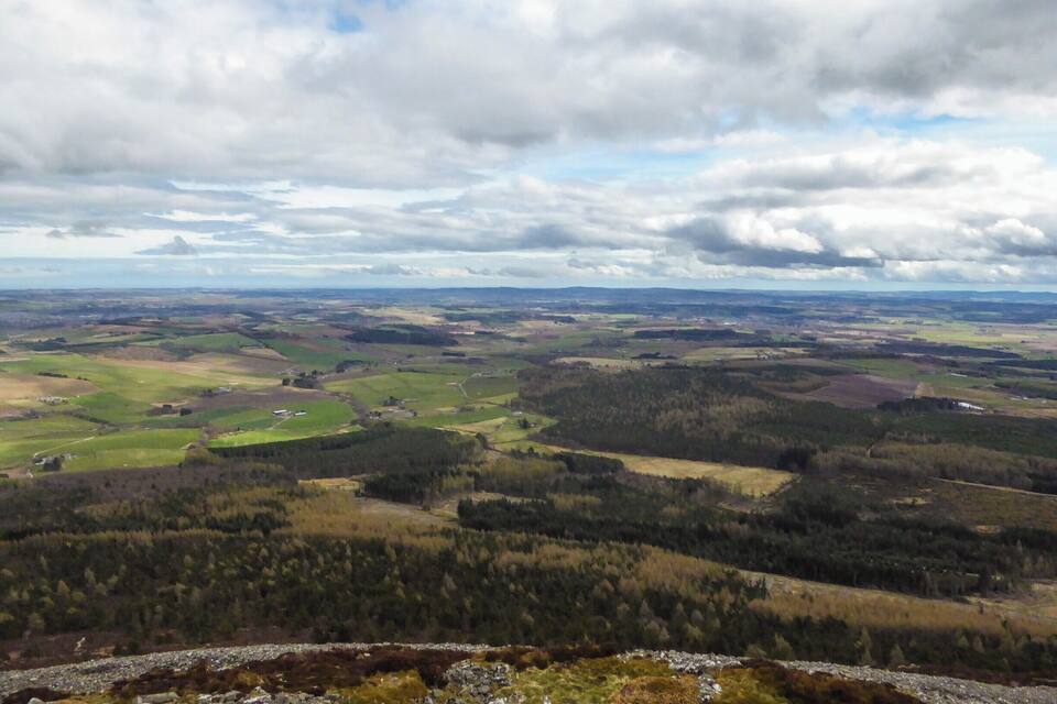 A panoramic view of a hilly landscape with scattered houses, dense forests, and a cloudy sky.