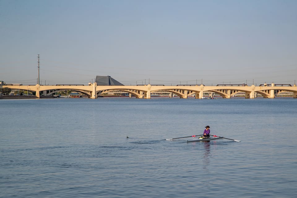 Une personne qui rame sur un bateau dans une rivière calme, avec un grand pont en arrière-plan.