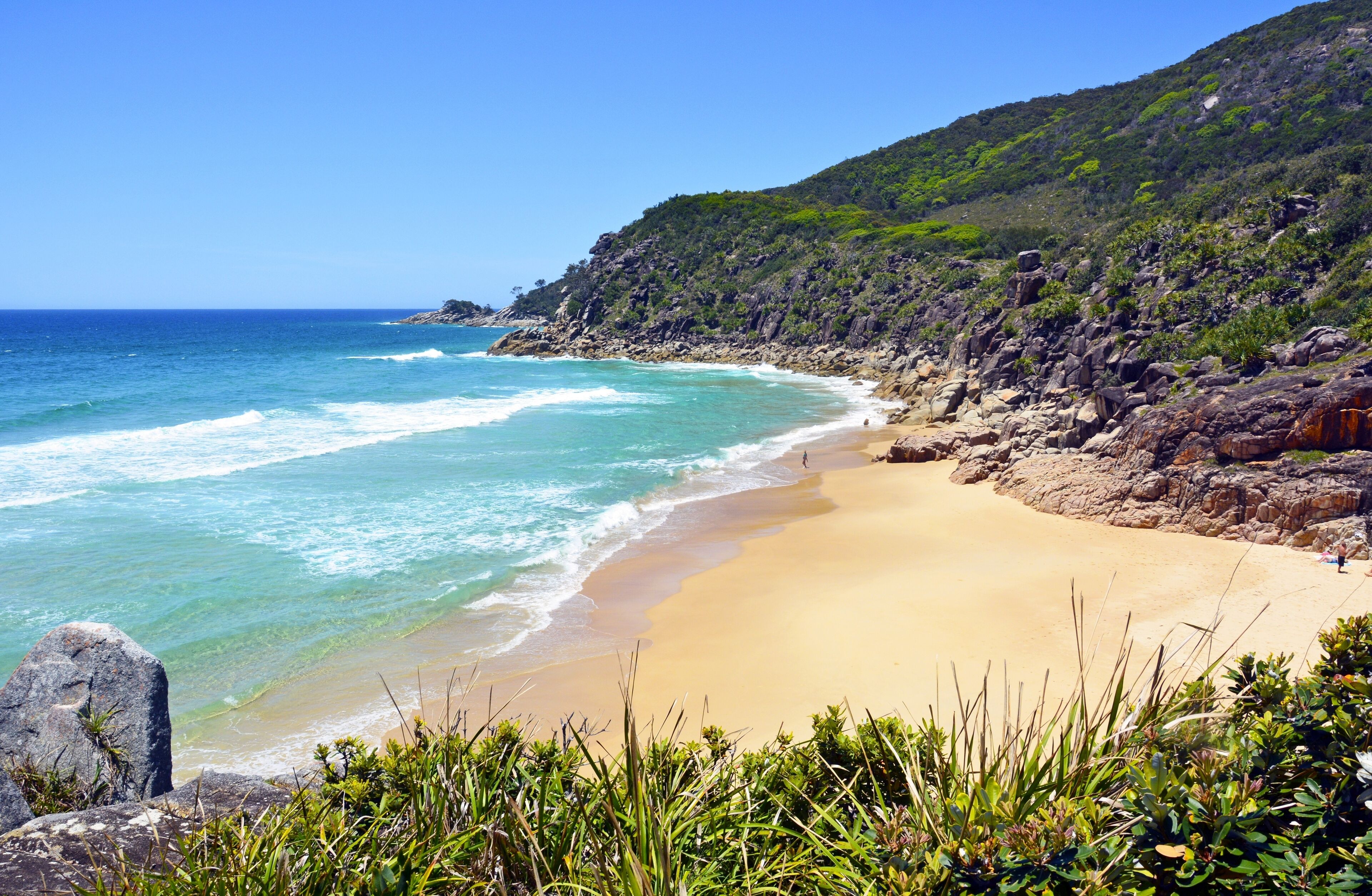 A coastal beach with clear turquoise waters, sandy shore, and a rocky hillside with greenery.