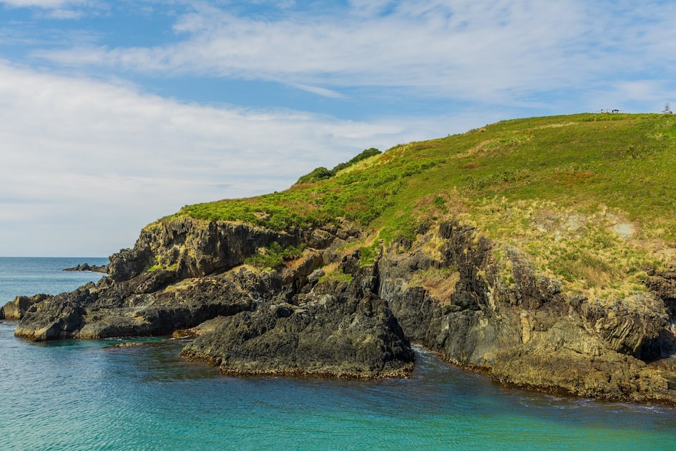 Une côte rocheuse bordée d’eaux turquoise et transparentes, et une colline verdoyante.