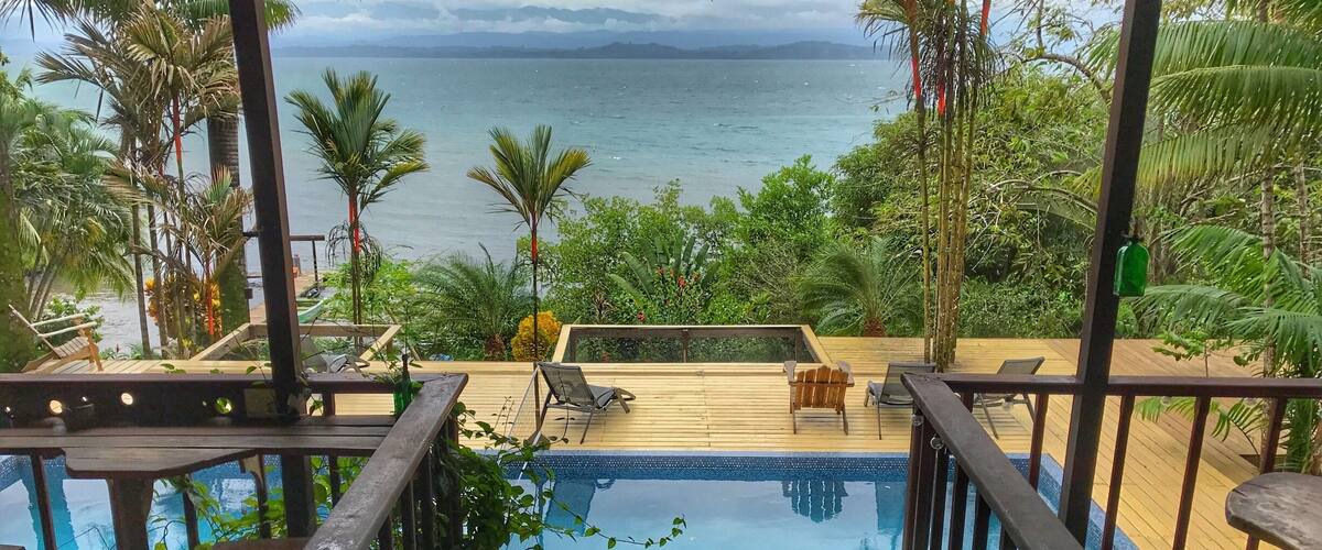 A pool with a view of the ocean, surrounded by palm trees and a wooden deck.