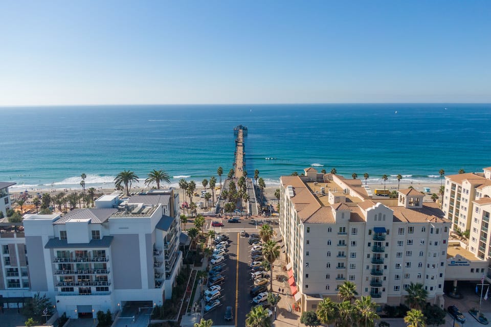 A coastal cityscape with a beach, palm trees, and a pier extending into the ocean.