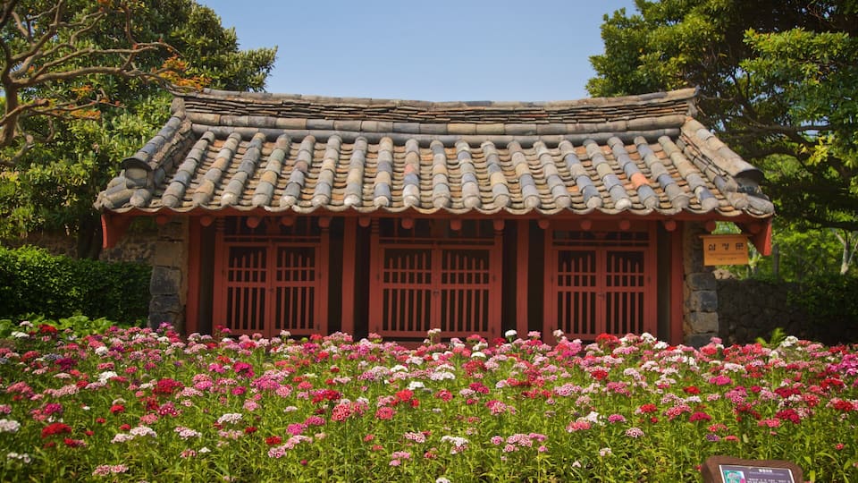 Un bâtiment traditionnel coréen, avec un toit recouvert de tuiles et des portes rouges, entouré d’un jardin où s’épanouissent des fleurs roses et blanches.