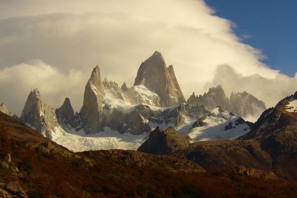 Una cordillera con picos destacados, laderas nevadas y un valle con vegetación rojiza.