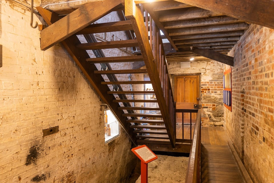 Un escalier en bois, dans un bâtiment de style rustique aux murs en briques apparentes, avec une porte en bois au sommet.