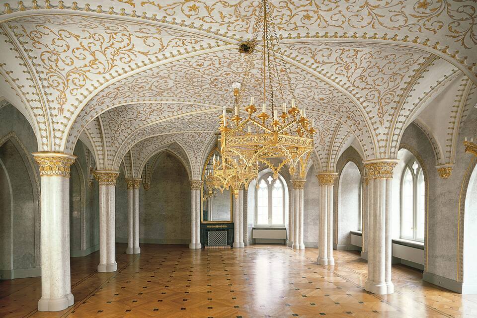 An ornate hall with a high ceiling, intricate gold detailing, and a large chandelier.