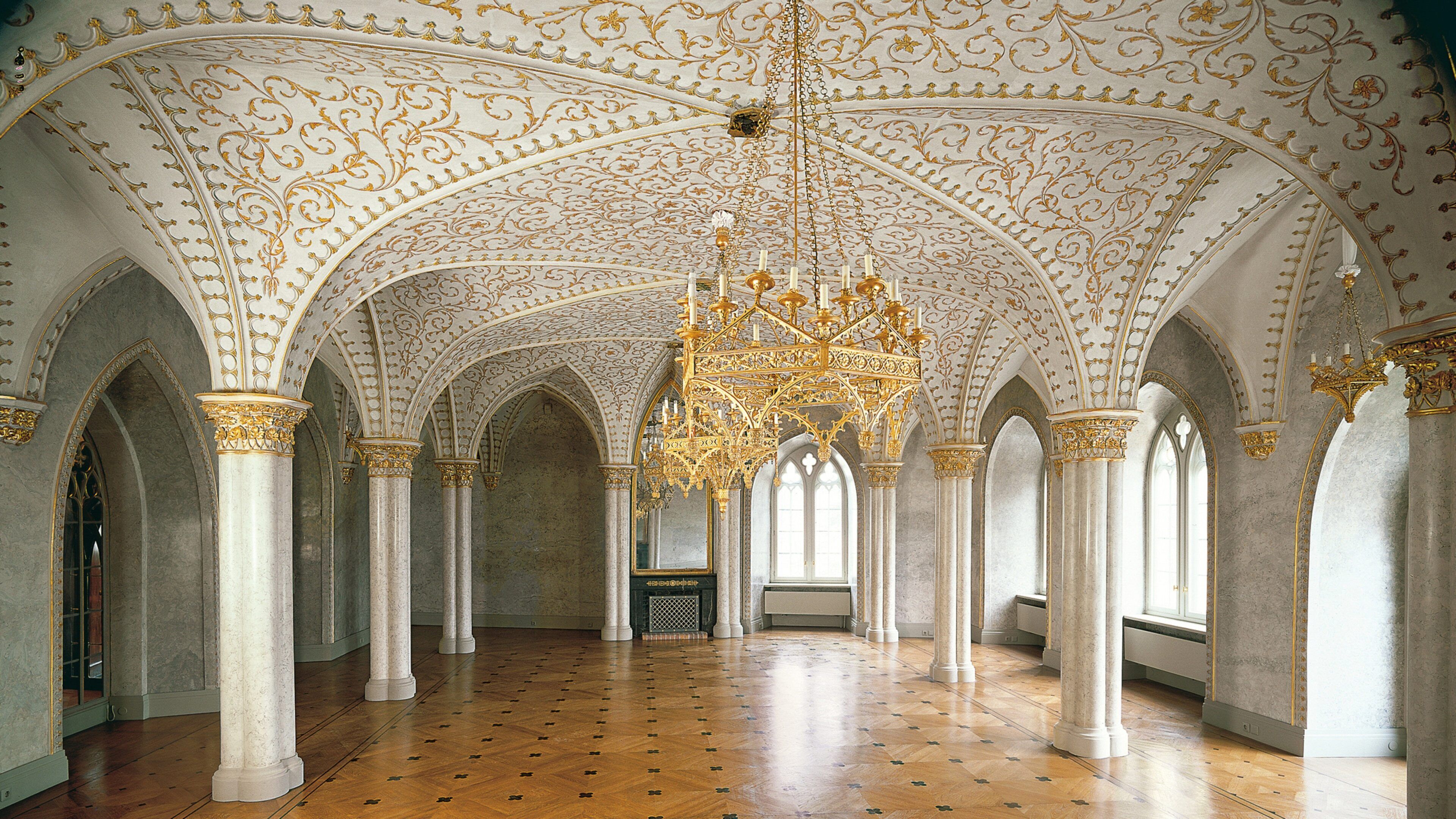 An ornate hall with a high ceiling, intricate gold detailing, and a large chandelier.