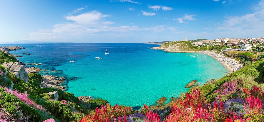 A coastal landscape with clear turquoise waters, rocky outcrops, and a beach with people.
