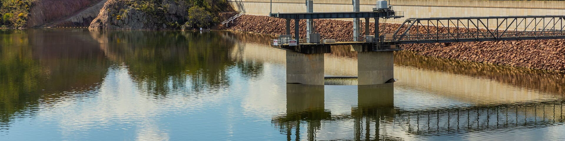 A dam with a bridge over a calm lake, surrounded by hills and trees.