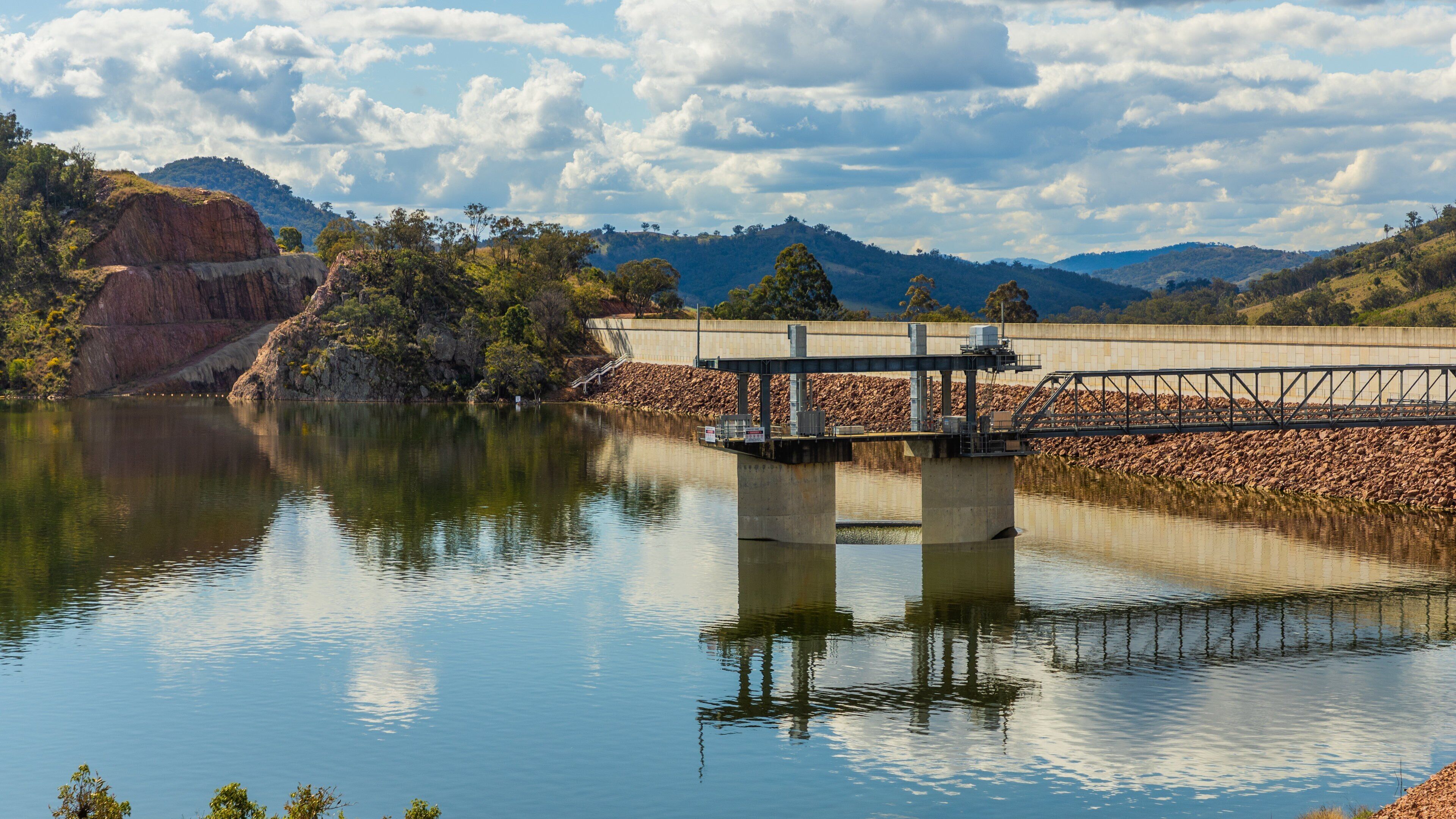 A dam with a bridge over a calm lake, surrounded by hills and trees.