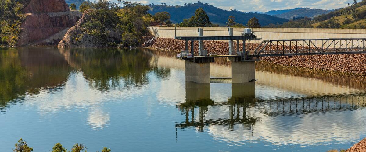 A dam with a bridge over a calm lake, surrounded by hills and trees.