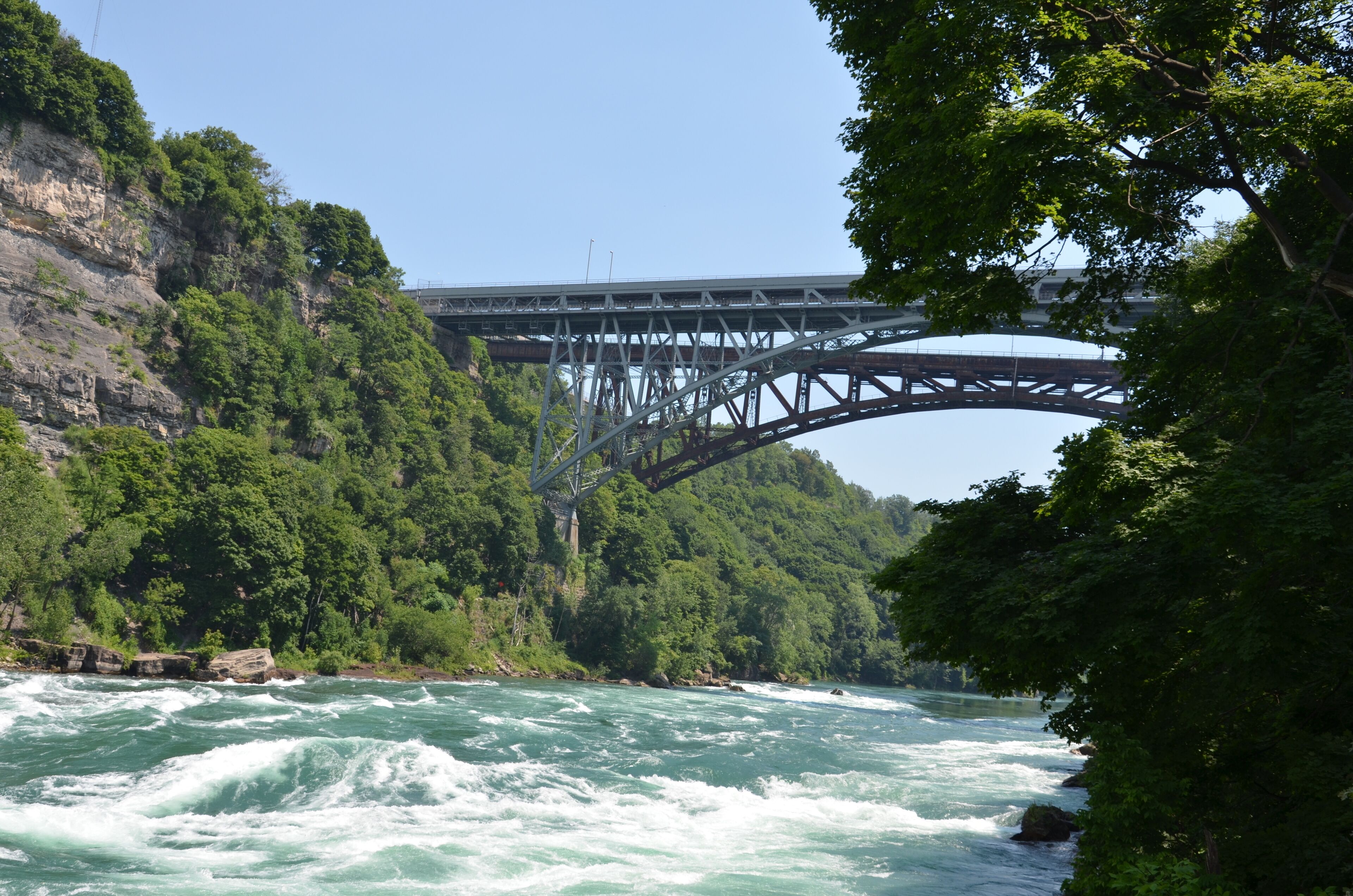 Bubbling rapids of the Niagara River flow over stones and rocks with bridge, forest and rock face in the background in sunny weather