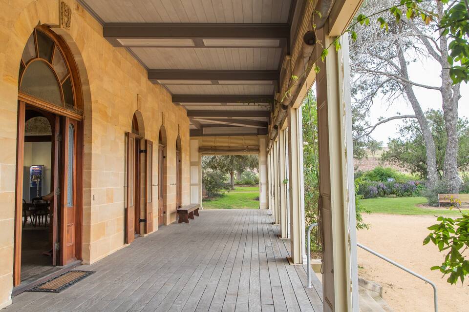 A covered porch with wooden doors and a bench, overlooking a grassy area with trees and a bench.