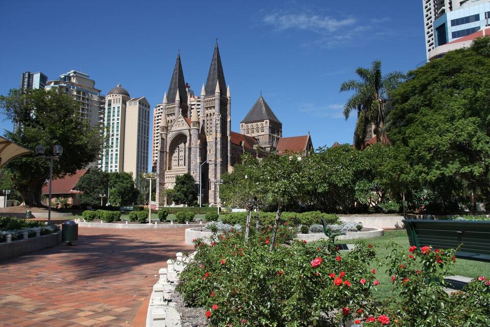 A historic church with twin spires, surrounded by a well-maintained garden with red roses, set against a backdrop of modern buildings and a clear blue sky.