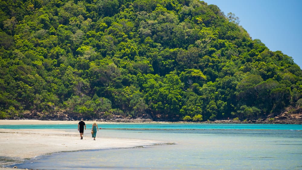 A beach with clear turquoise water and a forested hill in the background.