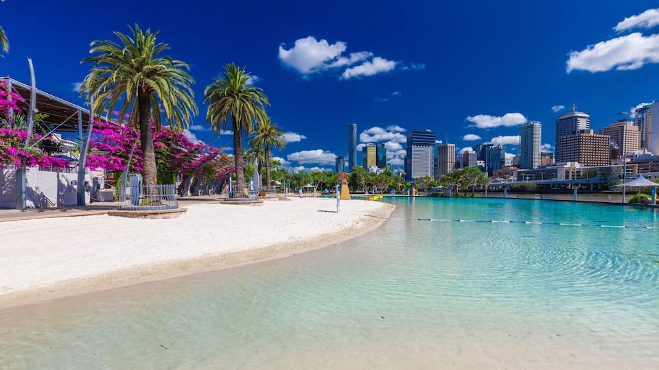 A beach with clear turquoise water, palm trees, and a city skyline in the background.