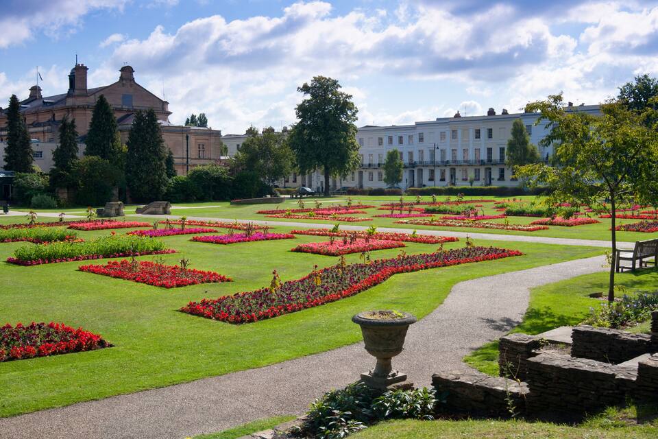 A formal garden with geometrically arranged flower beds and a pathway leading to a building with multiple chimneys.