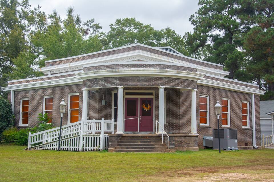 A brick building with a white porch, a red door, and a white fence.