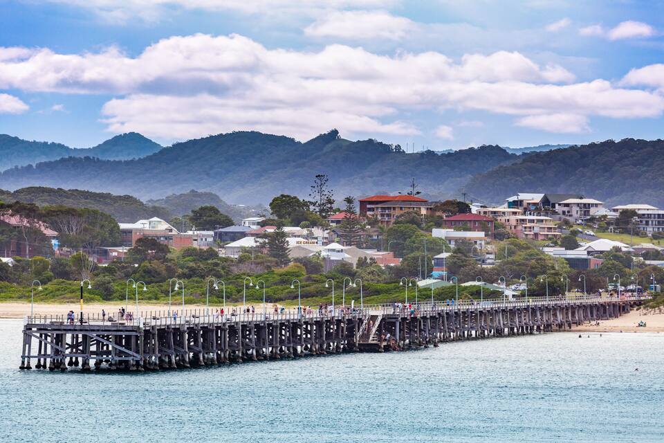 A pier extending into a calm sea with a beach and mountains in the background.