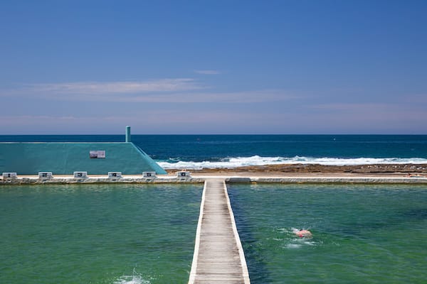 Une jetée s’avançant dans des eaux turquoise, bordée d’une côte rocheuse et surplombée d’un ciel bleu clair.