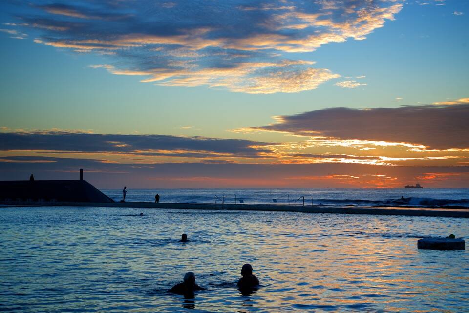 A sunset over a beach with people swimming and a ship in the distance.