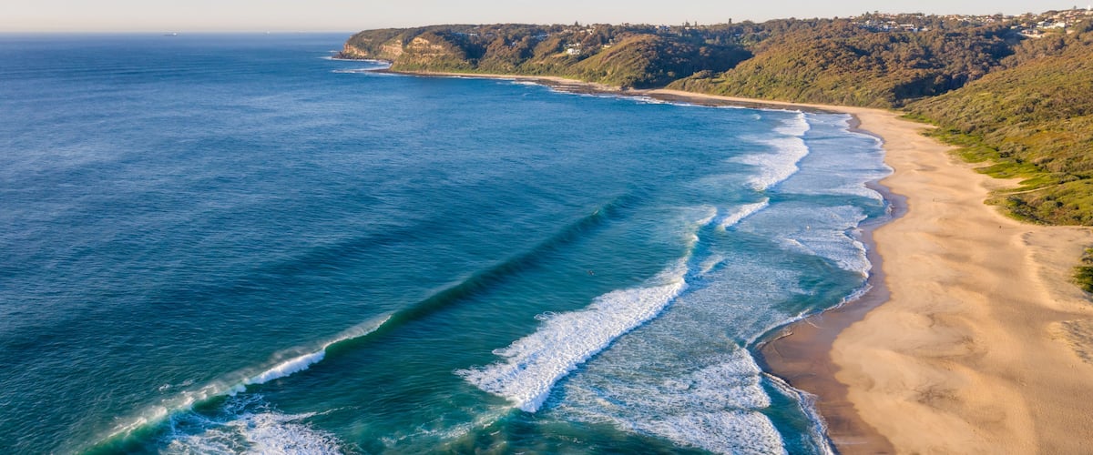 A coastal landscape with a sandy beach, ocean waves, and a hilly terrain with vegetation.