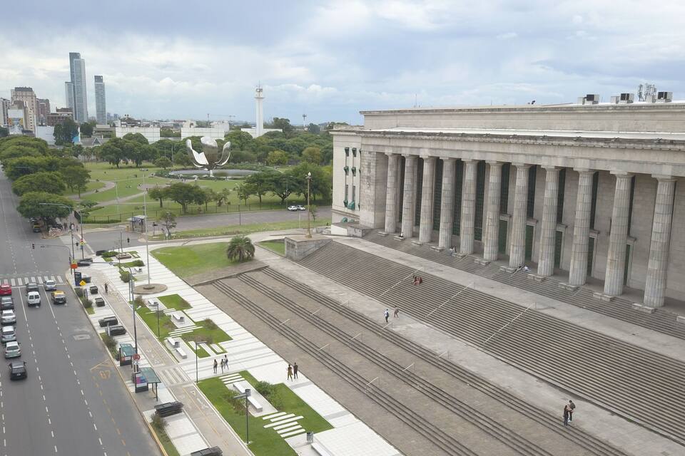 A cityscape with a large classical building with columns, a busy street, and a park with a sculpture.