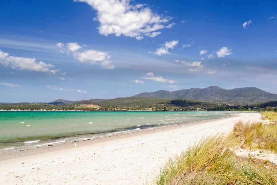 A beach with clear turquoise water, white sand, and green hills in the background.