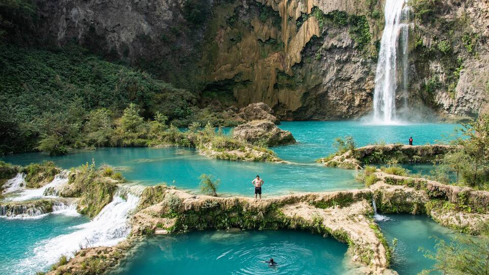 A waterfall cascading into a turquoise pool surrounded by rocky terrain and greenery.