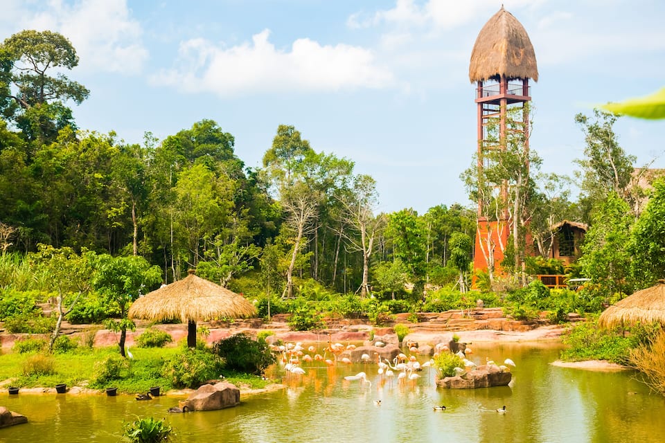 Üppige Landschaft mit einem Gewässer, Flamingos und einem hohen Turm mit einem Reetdach.