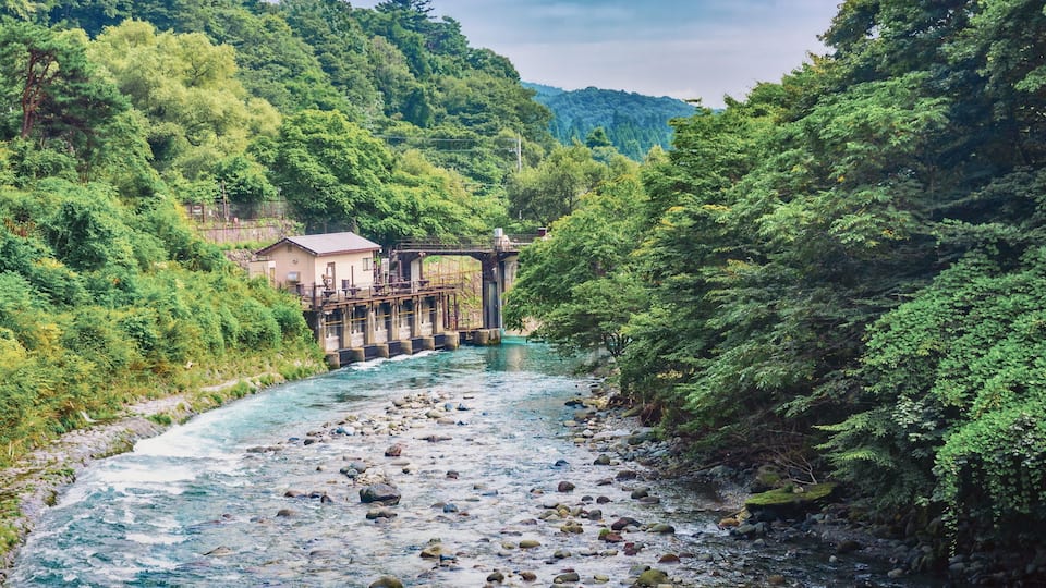 A river flowing through a forested area with a bridge and a small building.
