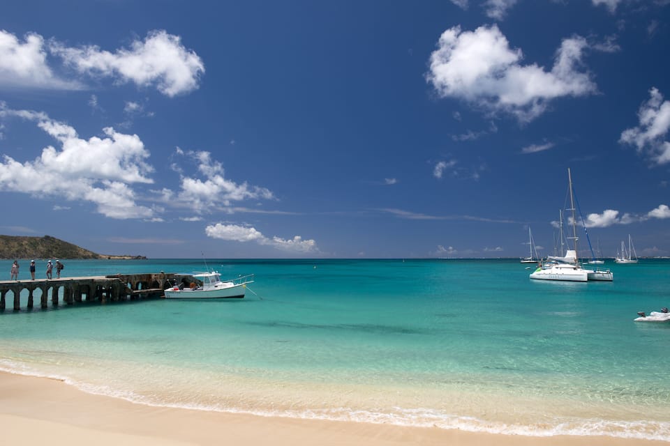 A beach with turquoise water, a pier, and sailboats.