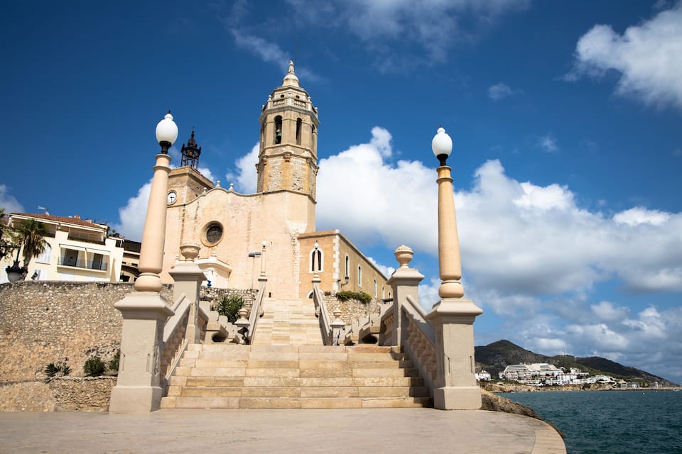 A historic church with a bell tower, steps leading up to it, and a coastal view in the background.