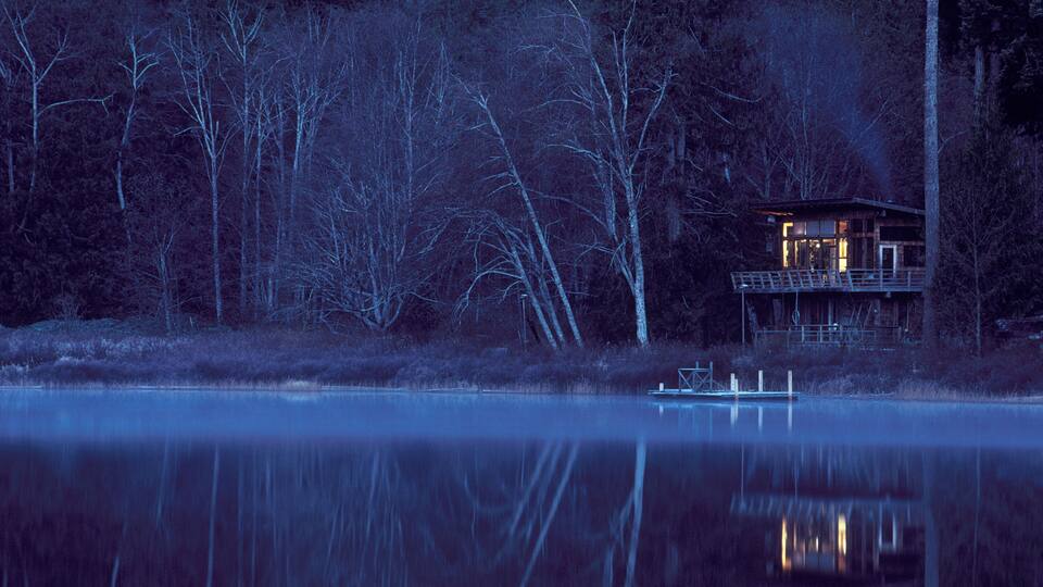 A cabin by a calm lake at dusk, surrounded by trees.