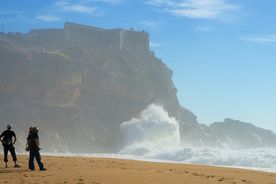 Una playa con olas rompiendo en la orilla, un acantilado rocoso con una estructura en la cima, y dos personas de pie sobre la arena.
