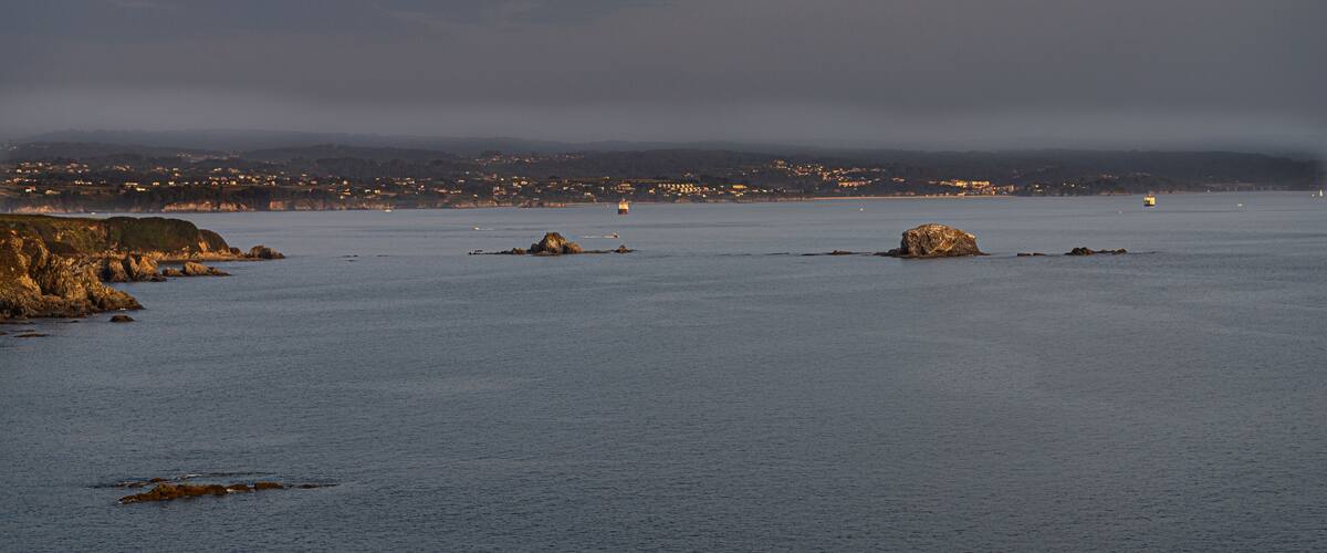 A coastal landscape with rocky outcrops and a distant cityscape under a twilight sky.
