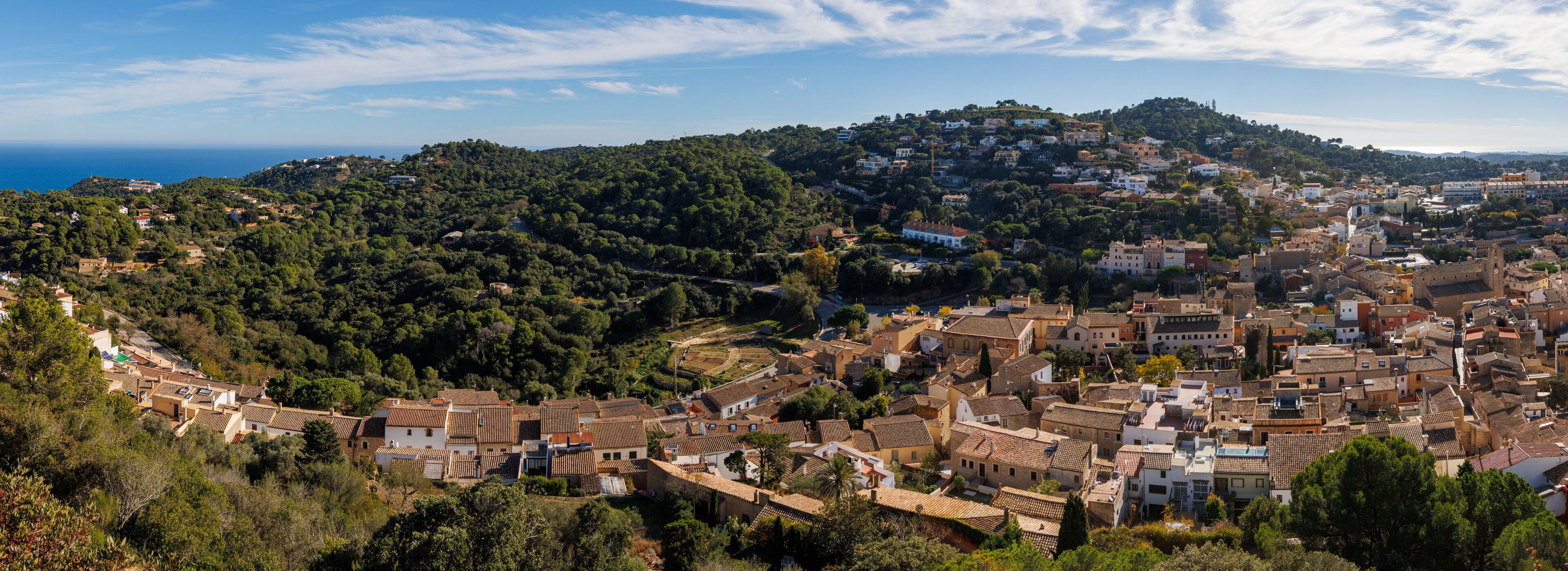 A coastal town with terracotta-roofed buildings, a hillside with dense greenery, and a clear view of the sea.