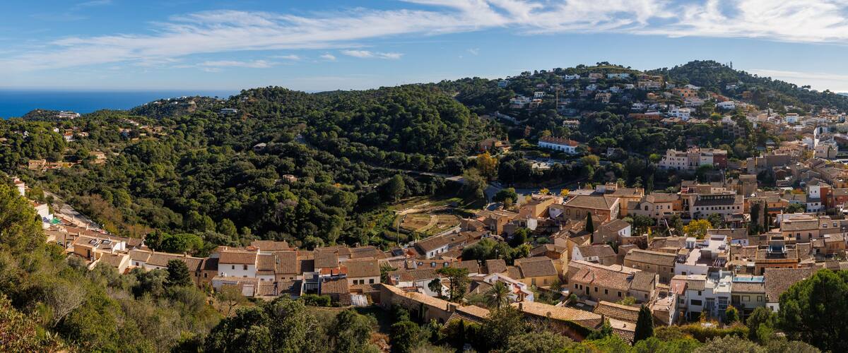 A coastal town with terracotta-roofed buildings, a hillside with dense greenery, and a clear view of the sea.